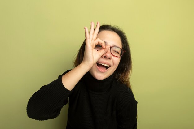 Giovane bella ragazza in un dolcevita nero e occhiali sorridente con la faccia felice che mostra segno giusto