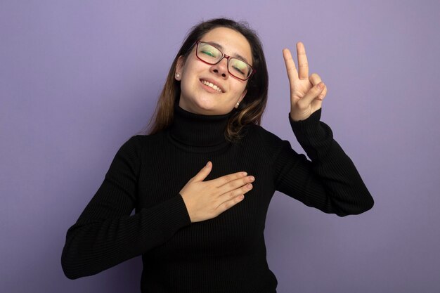 Giovane bella ragazza in un dolcevita nero e occhiali tenendo la mano sul petto che mostra v-segno sensazione di emozioni positive