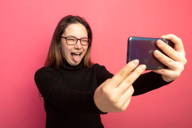 Giovane bella ragazza in un dolcevita nero e occhiali utilizza lo smartphone facendo selfie sorridente con la faccia felice che attacca fuori la lingua