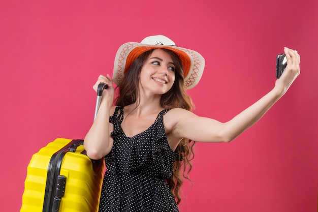 Giovane bella ragazza in vestito a pois in cappello estivo in piedi con la valigia guardando lo schermo del suo telefono cellulare sorridente allegramente prendendo selfie su sfondo rosa