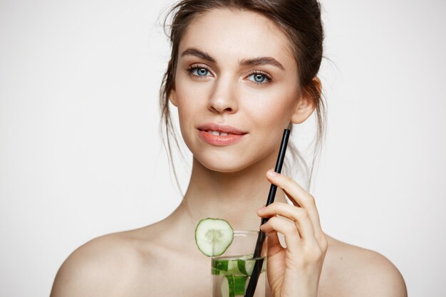 Giovane bella ragazza nuda con pelle pulita perfetta che sorride esaminando macchina fotografica che tiene bicchiere d'acqua con le fette del cetriolo sopra fondo bianco. Trattamento facciale.