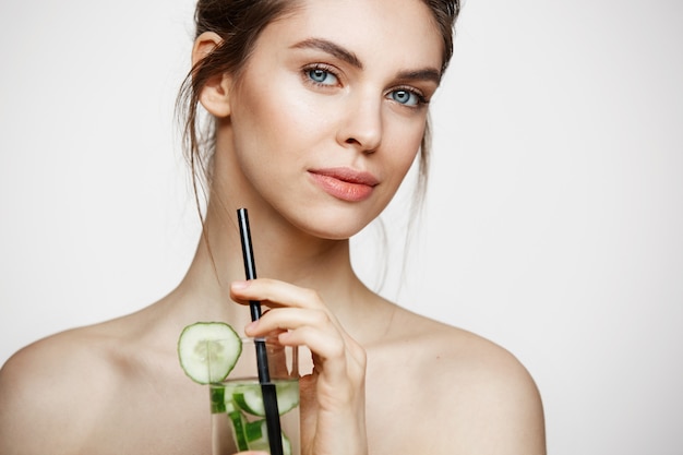 Giovane bella ragazza nuda con pelle pulita perfetta che sorride esaminando macchina fotografica che tiene bicchiere d'acqua con le fette del cetriolo sopra fondo bianco. Trattamento facciale.