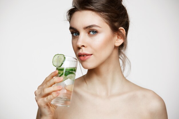 Giovane bella ragazza nuda con pelle pulita perfetta che sorride esaminando macchina fotografica che tiene bicchiere d'acqua con le fette del cetriolo sopra fondo bianco. Trattamento facciale.