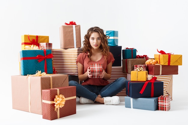 Giovane bella ragazza riccia che si siede sul pavimento fra i contenitori di regalo isolati