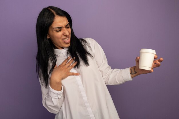 Giovane bella ragazza schizzinosa che indossa la maglietta bianca che tiene e che esamina la tazza di caffè che mette la mano sul cuore isolato sulla porpora