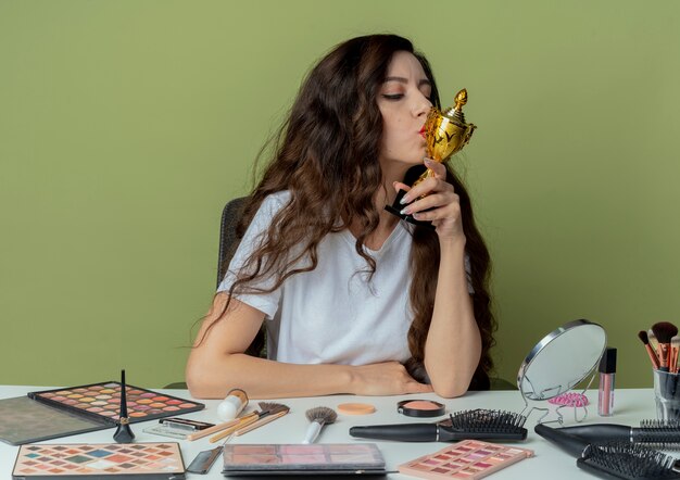 Giovane bella ragazza seduta al tavolo di trucco con strumenti di trucco tenendo e baciando la tazza del vincitore isolato su sfondo verde oliva