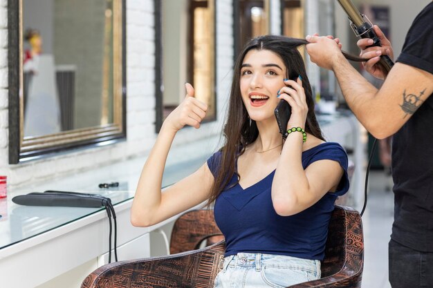 Giovane bella ragazza seduta dal barbiere e gesto con il pollice in su mentre parla al telefono Foto di alta qualità