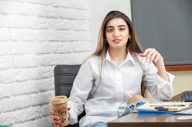 Giovane bella ragazza seduta dietro la scrivania e in possesso di una tazza di caffè Foto di alta qualità