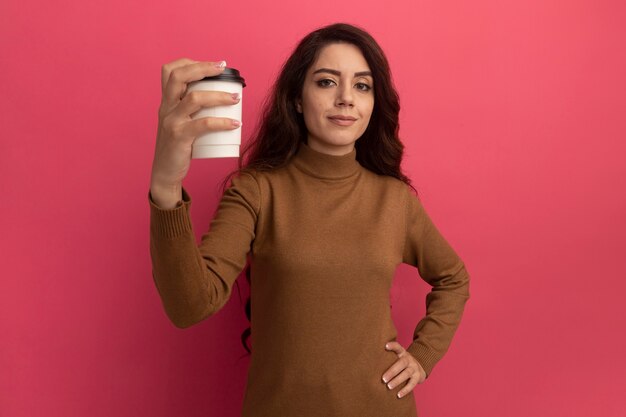 Giovane bella ragazza sicura che porta il maglione a collo alto che tiene tazza di caffè isolata sulla parete rosa