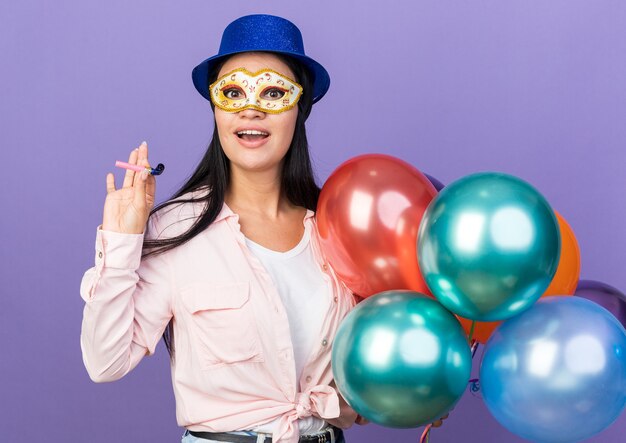 Giovane bella ragazza sorpresa che indossa cappello da festa e maschera per gli occhi in maschera che tiene palloncini con fischietto da festa