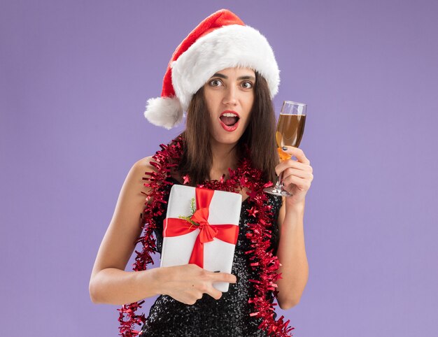 Giovane bella ragazza sorpresa che indossa il cappello di Natale con la ghirlanda sul collo che tiene il contenitore di regalo con un bicchiere di champagne isolato su sfondo viola