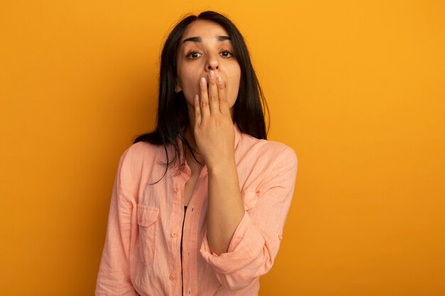 Giovane bella ragazza sorpresa che indossa la bocca coperta di maglietta rosa con la mano isolata su colore giallo con lo spazio della copia