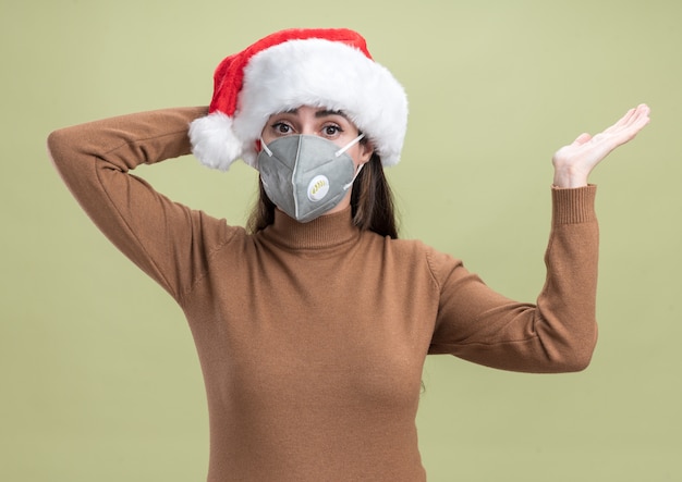 Giovane bella ragazza sorpresa che porta il cappello di natale con i punti medici della mascherina con la mano a lato che mette la mano sulla testa isolata su fondo verde oliva