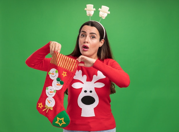 Giovane bella ragazza sorpresa che porta il maglione di natale con il cerchio di capelli di natale che tiene i calzini di natale isolati su fondo verde