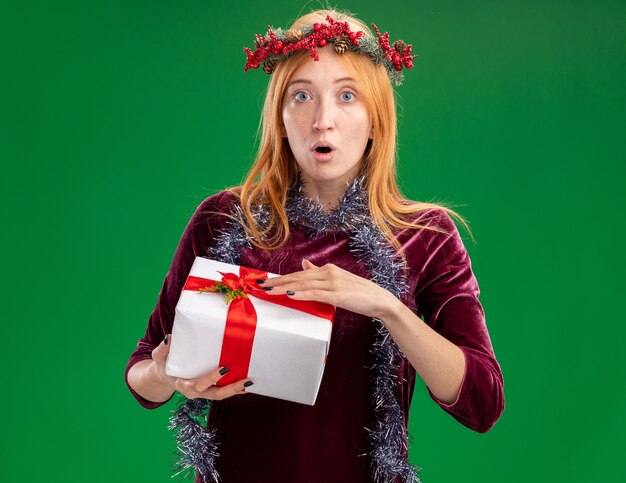 Giovane bella ragazza sorpresa che porta vestito rosso con la corona e la ghirlanda sul collo che tiene il contenitore di regalo isolato su fondo verde