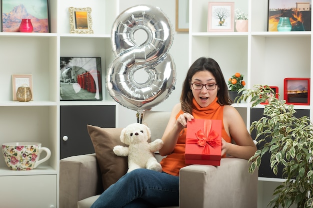 Giovane bella ragazza sorpresa in occhiali ottici che si apre e guarda la confezione regalo seduta sulla poltrona in soggiorno a marzo giornata internazionale della donna