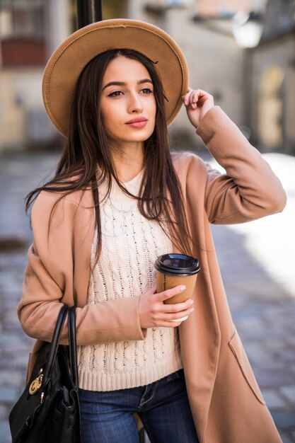 Giovane bella signora che cammina lungo la strada con borsetta e tazza di caffè.
