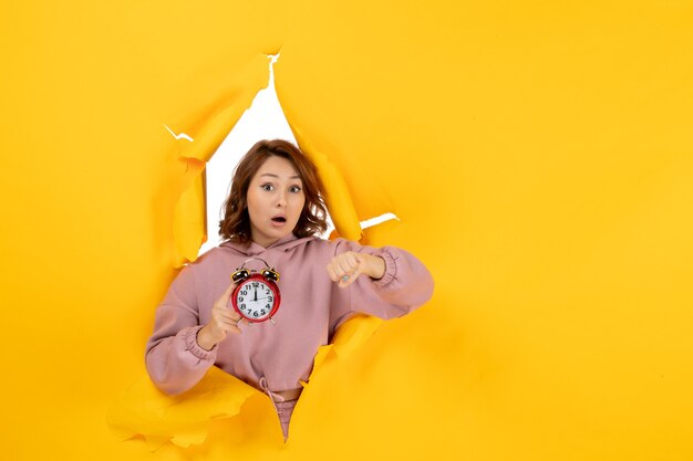 Giovane bella signora sorpresa che tiene l'orologio e controlla il suo tempo su uno sfondo giallo strappato