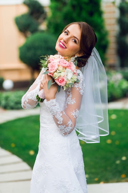 Giovane bella sposa felice in vestito da sposa elegante bianco con il mazzo che posa all'aperto nel parco.