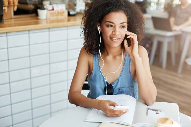 Giovane bella studentessa africana che ascolta la musica in seduta sorridente delle cuffie alla tavola con i libri al caffè.