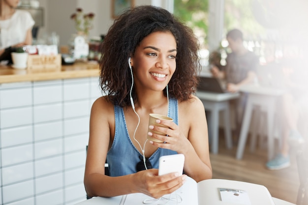 Giovane bella studentessa africana che ascolta la musica in seduta sorridente delle cuffie alla tavola con i libri al caffè.