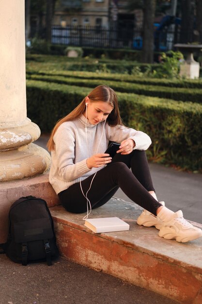 Giovane bella studentessa alla moda in maglione accogliente che usa sognante il cellulare durante la pausa di studio all'aperto