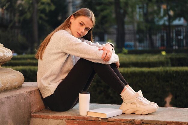 Giovane bella studentessa alla moda in un maglione accogliente che guarda premurosamente a porte chiuse mentre studia all'aperto