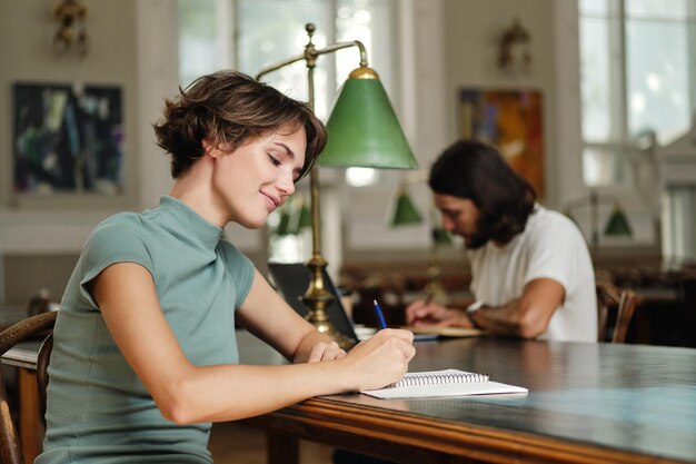 Giovane bella studentessa che prende felicemente appunti mentre studia nella biblioteca dell'università