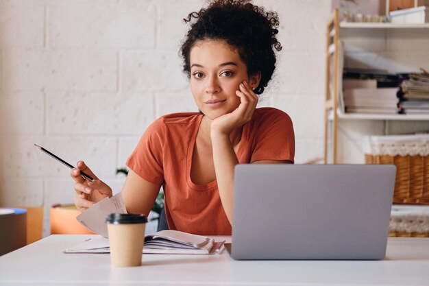 Giovane bella studentessa con i capelli ricci scuri seduta al tavolo con laptop e tazza di caffè per andare appoggiandosi sulla mano guardando sognante in camera con la matita mentre studiava a casa accogliente