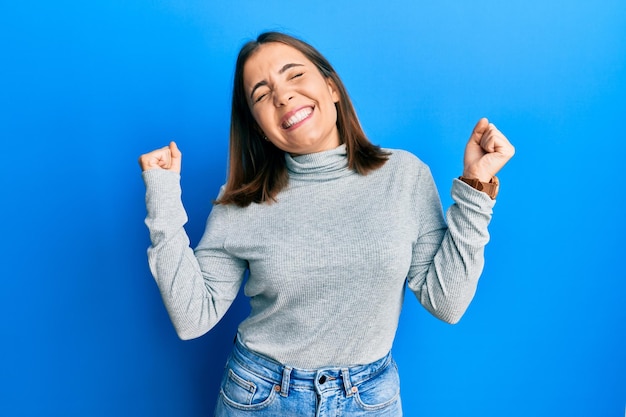 Giovane bellissima donna che indossa un maglione a collo alto molto felice ed eccitato facendo il gesto del vincitore con le braccia alzate sorridendo e urlando per il successo concetto di celebrazione