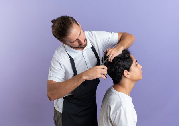 Giovane bello barbiere che indossa uniforme facendo taglio di capelli per il giovane cliente isolato su sfondo viola con copia spazio