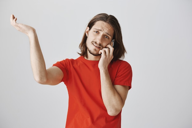 Giovane bello indeciso che parla sul telefono e che scrolla le spalle all'oscuro