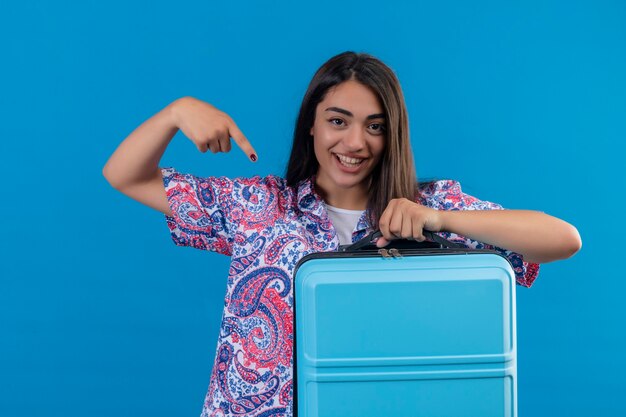 Giovane bello viaggiatore donna che tiene la valigia che punta con il dito indice ad esso guardando fiducioso positivo e sorridente felice allegramente pronto a viaggiare in piedi su sfondo blu