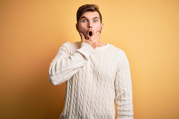 Giovane biondo con la barba e gli occhi azzurri che indossa un maglione bianco su sfondo giallo che sembra affascinato dall'incredulità sorpresa e dall'espressione stupita con le mani sul mento