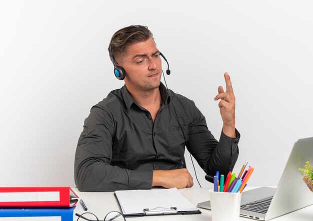 Giovane biondo infastidito lavoratore di ufficio uomo sulle cuffie si siede alla scrivania con strumenti di ufficio guardando il laptop con la mano sollevata isolati su sfondo bianco con spazio di copia