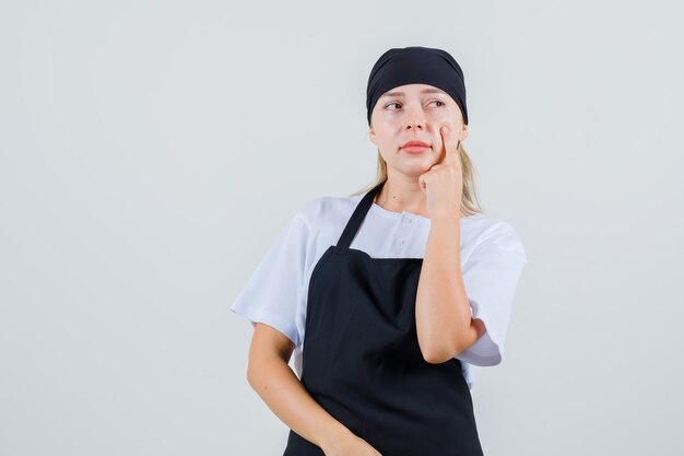 Giovane cameriera che guarda lontano con il dito sulla guancia in uniforme e grembiule