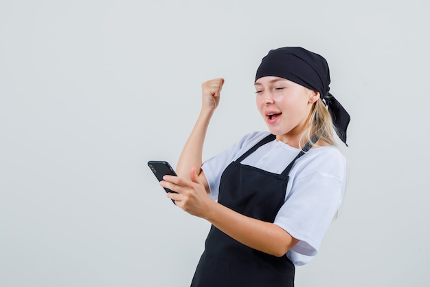 Giovane cameriera guardando il cellulare in uniforme e grembiule e guardando felice