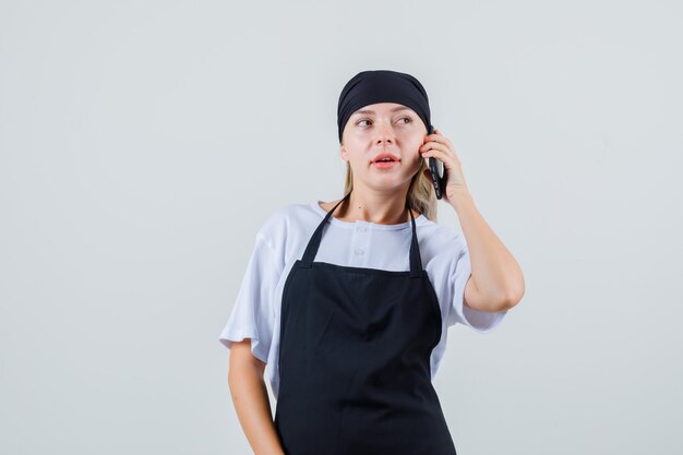 Giovane cameriera in uniforme e grembiule che guarda lontano mentre parla al telefono cellulare
