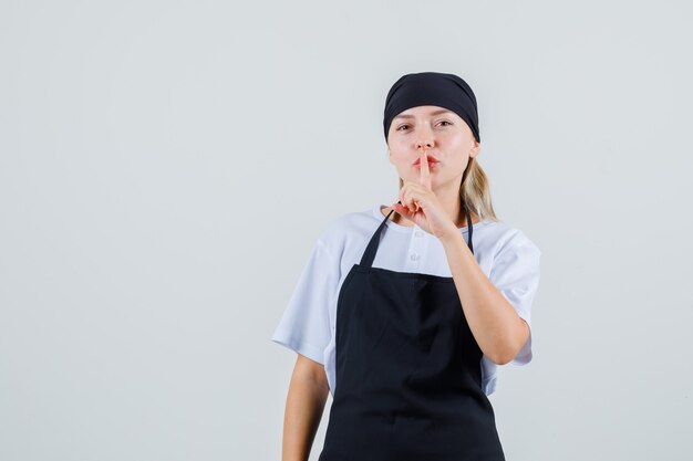 Giovane cameriera in uniforme e grembiule che mostra gesto di silenzio e che osserva attento