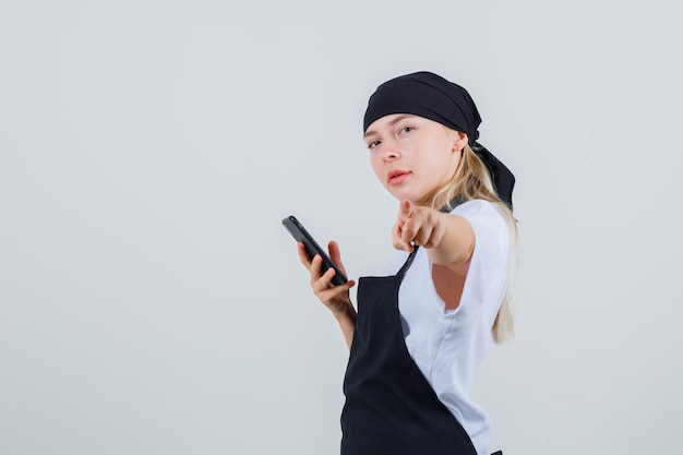 Giovane cameriera in uniforme e grembiule che punta mentre si tiene lo smartphone.