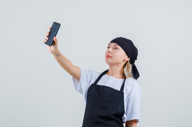 Giovane cameriera in uniforme e grembiule tenendo selfie sul cellulare