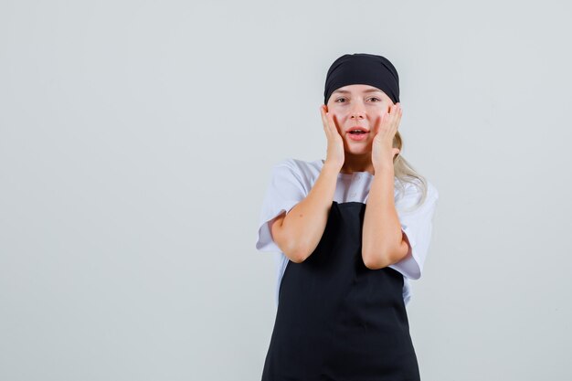 Giovane cameriera tenendo le mani sulle guance in uniforme e grembiule e guardando stupito