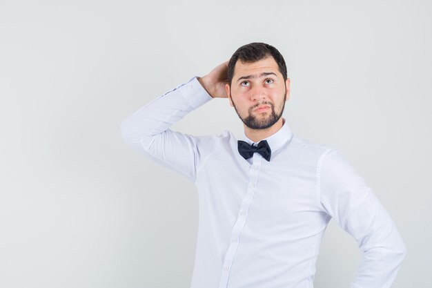 Giovane cameriere che osserva in su con la mano dietro la testa in camicia bianca e che sembra pensieroso. vista frontale.