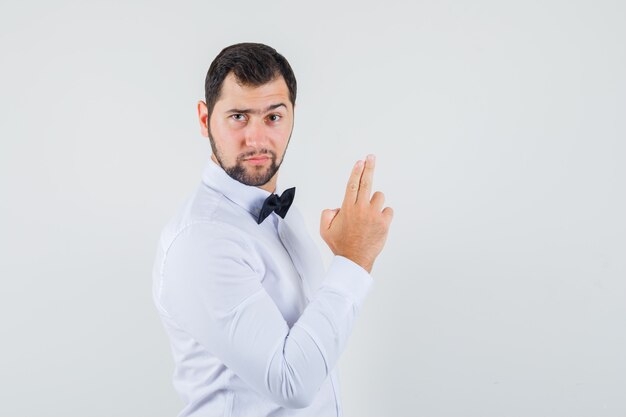 Giovane cameriere in camicia bianca che fa il segno della pistola del dito e che sembra fiducioso, vista frontale.