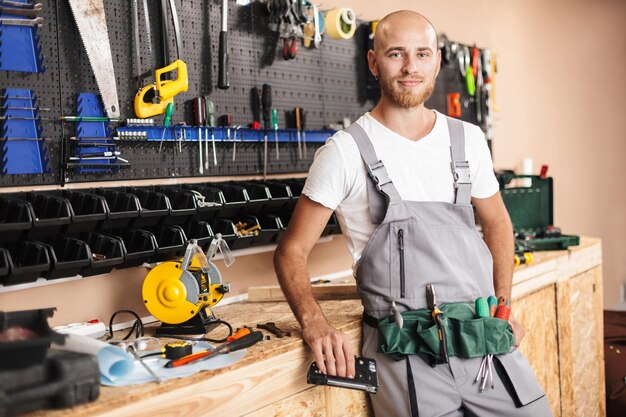 Giovane caposquadra sorridente in abiti da lavoro che tiene in mano la cucitrice meccanica guardando sognante nella fotocamera con supporto di diversi strumenti sullo sfondo in officina