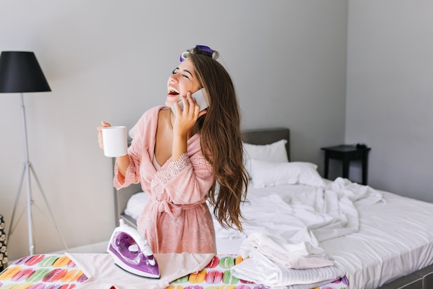 Giovane casalinga con i capelli lunghi in accappatoio rosa con bigodino sulla testa a casa a stirare i vestiti. Parla al telefono, ride con gli occhi chiusi.