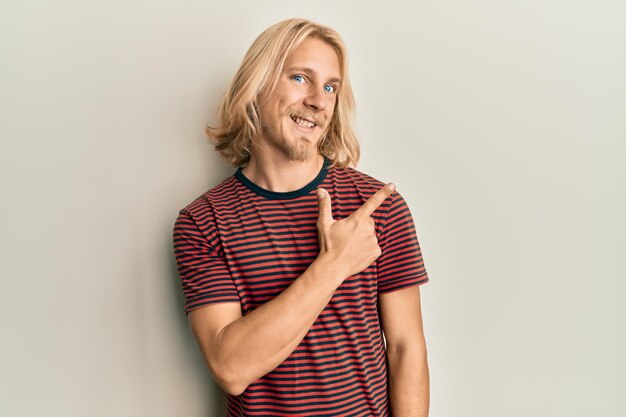 Giovane caucasico con i capelli lunghi che indossa una maglietta a righe casual che sorride allegro indicando con la mano e il dito verso l'alto