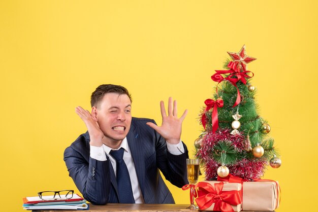 giovane che ascolta qualcosa seduto al tavolo vicino all'albero di Natale e regali su giallo