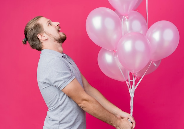 Giovane che celebra la festa di compleanno tenendo un mazzo di palloncini in piedi lateralmente cercando felice ed eccitato oltre il muro rosa