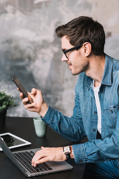 Giovane che digita sul computer portatile e che esamina cellulare